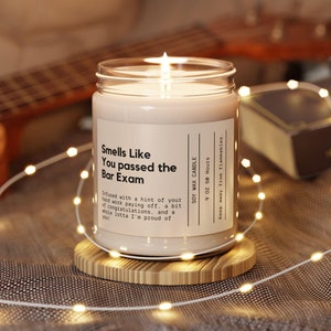 Puede incluir: Un tarro de vela de vidrio transparente con una vela de cera de soja beige en su interior. La vela tiene una etiqueta que dice "Smells Like You Passed the Bar Exam" e incluye el texto "Soy Wax Candle, 9 oz, 50 Hours, Keep away from flammables".