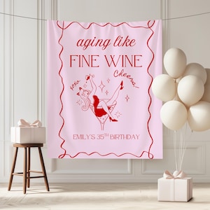 May include: A pink banner with red text and a red illustration of a woman in a martini glass. The text reads "aging like FINE WINE Cheers, xoxo. EMILY'S 35TH BIRTHDAY". White balloons and a gift box are also visible.