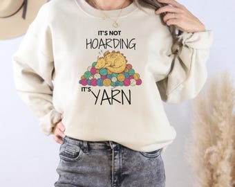 May include: Beige sweatshirt with the text "IT'S NOT HOARDING IT'S YARN". The graphic shows a sleeping dragon on a pile of colorful yarn balls. A fun, cozy top for knitters and crafters.