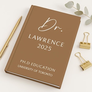 Op de afbeelding: Een bruin hardcover-dagboek met de naam "Dr. Lawrence 2025" in witte letters. Het dagboek bevat ook de tekst "PH.D EDUCATION UNIVERSITY OF TORONTO." Een gouden pen en twee gouden paperclips zijn ook zichtbaar.