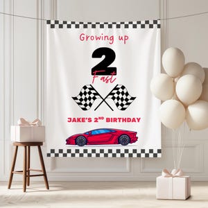 May include: White banner with a black and white checkered border, featuring the text "Growing up 2 Fast" and "Jake's 2nd Birthday." Includes a red sports car graphic, checkered flags, and white balloons. A gift box sits on a wooden stool.