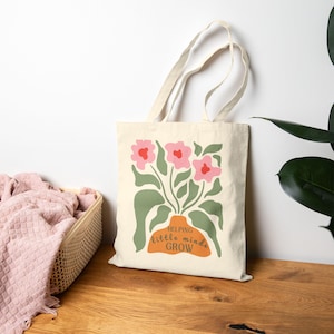 Puede incluir: Un bolso tote crema con un diseño floral. Presenta flores rosas, hojas verdes y un jarrón naranja con el texto "Helping Little Minds Grow". El bolso tiene asas color crema. El diseño es una ilustración sencilla y colorida.