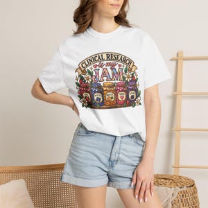 Peut inclure: Un t-shirt blanc à manches courtes avec un graphique coloré représentant cinq bocaux en verre sur une étagère. Le texte indique CLINICAL RESEARCH is my JAM. Chaque bocal est étiqueté avec des mots comme DATA, PROTOCOLS, ANALYSIS, PATIENTS et OUTCOMES.