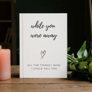 Może przedstawiać: Biały notatnik z napisem "while you were away" i "All the things I wish I could tell you" czarnym pismem. Obok na drewnianej powierzchni stoi zapalona brzoskwiniowa świeca i pomarańczowa róża z zielenią.