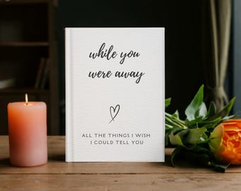 May include: A white journal with the text "while you were away" and "All the things I wish I could tell you" in black script. A lit peach-colored candle and an orange rose with greenery sit nearby on a wooden surface.