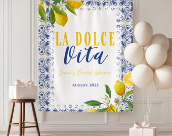 May include: A white banner with blue and white floral border and the text "LA DOLCE VITA" in yellow and blue. The banner includes lemon illustrations and the text "Jenni's Bridal Shower" and "AUGUST, 2025". White balloons and a gift box are in the background.