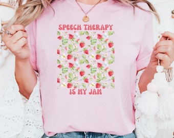 May include: Pink t-shirt with a strawberry pattern and the text "SPEECH THERAPY IS MY JAM" in a retro font. The design features red strawberries, green leaves, and white flowers.