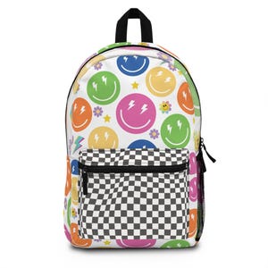 May include: A white backpack with a checkered pattern on the front pocket. The main body of the backpack is covered in a pattern of colorful smiley faces with lightning bolts and flowers.