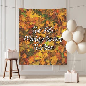 Puede incluir: Un telón de fondo con temática otoñal con el texto "The Fall Maddy turned Thirteen". El telón de fondo presenta una vibrante exhibición de hojas de otoño en tonos amarillos, naranjas y rojos. También hay globos blancos y una caja de regalo.