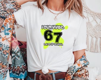 Può includere: T-shirt bianco con la scritta "I SURVIVED 67 DAYS OF SCHOOL" in nero e verde. La maglietta ha un girocollo e maniche corte. La maglietta è abbinata a jeans blu e una cintura marrone.