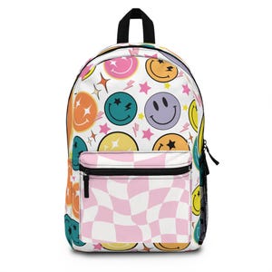 May include: A white backpack with a pink and white checkered pattern on the front pocket. The backpack is covered in a pattern of colorful smiley faces, stars, and lightning bolts.