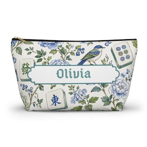 May include: A zipped pouch with a floral and bird design. The pouch has a white background with blue floral patterns, a blue bird, and the name "Olivia" in teal. Mahjong tiles are also visible.