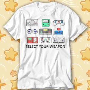 May include: White t-shirt featuring a grid of colorful retro video game consoles and controllers. The text "SELECT YOUR WEAPON" is printed below the graphics. The design includes a variety of game consoles in a playful, cartoon style.