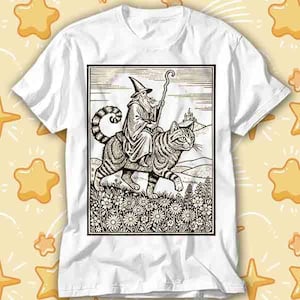 May include: White t-shirt with a black and white illustration of a wizard riding a striped cat. The wizard wears a pointed hat and holds a staff. The cat has a curled tail and is walking through a field of flowers.