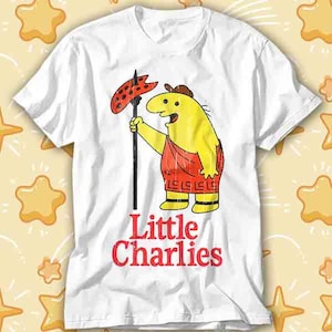 May include: White t-shirt featuring a cartoon character with a yellow body, red shorts, and a brown hat. The character holds a black spear with a red, dotted object. The words "Little Charlies" are printed in red.