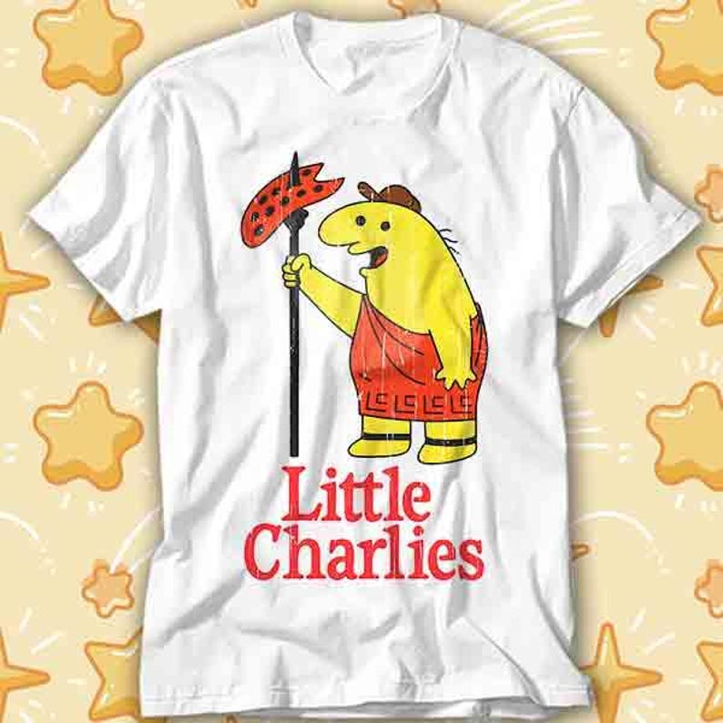 May include: White t-shirt featuring a cartoon character with a yellow body, red shorts, and a brown hat. The character holds a black spear with a red, dotted object. The words "Little Charlies" are printed in red.