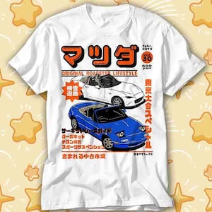 Puede incluir: Camiseta blanca con un gráfico de dos coches Mazda roadster, uno blanco y otro azul, y texto japonés. El diseño incluye las palabras "ORIGINAL ROADSTER LIFESTYLE" y tiene un estilo de revista vintage.