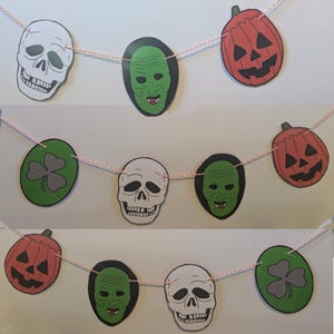 May include: A Halloween banner featuring alternating green shamrocks, white skulls, green monster faces, and orange pumpkins. The banner is strung with pink thread.