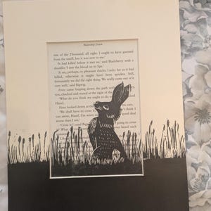 May include: A framed black and white print featuring a rabbit illustration set against a backdrop of text from a book. The artwork is mounted on a cream-colored mat and framed in a larger white frame, creating a layered effect. The print depicts a rabbit in a field.