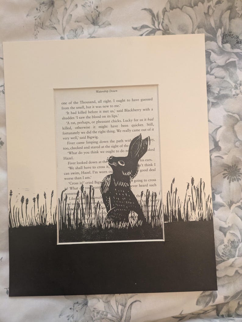 May include: A framed black and white print featuring a rabbit illustration set against a backdrop of text from a book. The artwork is mounted on a cream-colored mat and framed in a larger white frame, creating a layered effect. The print depicts a rabbit in a field.