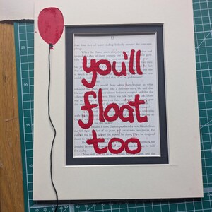 Könnte beinhalten: Ein gerahmtes Kunstwerk mit einem roten Ballon und den Worten "you'll float too" in roter Farbe auf einer Buchseite. Der Ballon ist mit schwarzem Faden an der Seite befestigt.