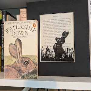 May include: A bookshelf featuring a copy of "Watership Down" by Richard Adams, with a rabbit illustration on the cover. Beside it is a framed black and white print of a rabbit in tall grass. The books are on a white shelf.