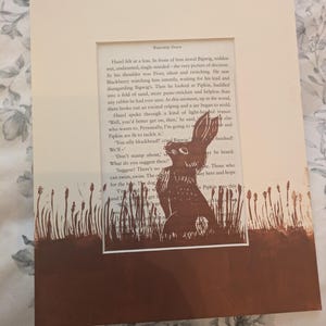 May include: A framed print featuring a brown illustration of a rabbit in a field of tall grass. The artwork is set against a backdrop of text from a book page. The frame is a neutral cream color, creating a rustic aesthetic.
