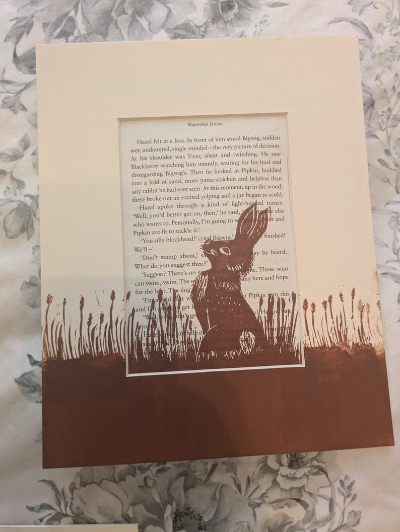 May include: A framed print featuring a brown illustration of a rabbit in a field of tall grass. The artwork is set against a backdrop of text from a book page. The frame is a neutral cream color, creating a rustic aesthetic.