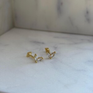 May include: A pair of gold earrings with small, clear, marquise-shaped stones. The earrings are set on a small, square post.