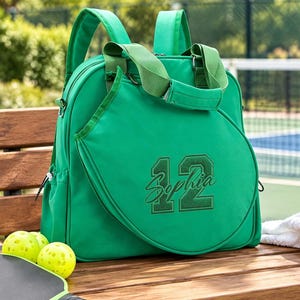 May include: A bright green pickleball backpack featuring a dedicated front compartment for a paddle, embroidered with the number 12 and the name Sophia in a matching green thread. The bag includes adjustable shoulder straps and top carry handles.