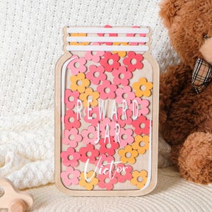 Peut inclure: Un pot à récompenses en bois en forme de bocal, rempli de découpes de fleurs roses et jaunes. Les mots "REWARD JAR" et le nom "Victor" sont affichés. Une voiture jouet en bois et un ours en peluche sont en arrière-plan.