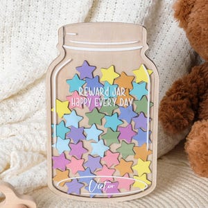 Peut inclure: Pot à récompenses en bois en forme de bocal, rempli d'étoiles colorées. Le bocal porte l'inscription "REWARD JAR HAPPY EVERY DAY" et un nom en bas. Une voiture jouet en bois et un ours en peluche sont en arrière-plan.