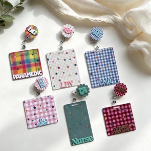 May include: Six rectangular badge reels featuring various patterns including plaid, gingham, and solid colours with star and moon accents. The reels display text labels such as PARAMEDIC, LPN, CMA, ER NURSE, Nurse, and PHYSICAL THERAPIST in raised lettering.