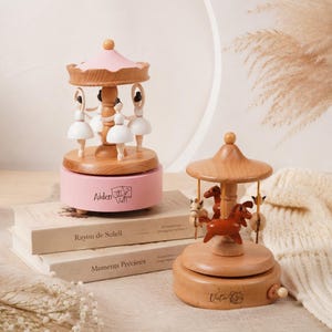 May include: Two wooden musical carousels. The left carousel has a pink roof and base with the name Aiden and a small bear graphic. It features four white-clad dancers. The right carousel is natural wood with brown horses and the name Victor.