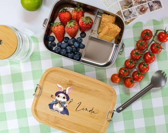 May include: A stainless steel lunch box with three compartments, filled with strawberries, blueberries, and a few pieces of bread. The lunch box is next to a bamboo lunch box with a white rabbit wearing a blue cape and a hat, and the name "Linda" written on it.