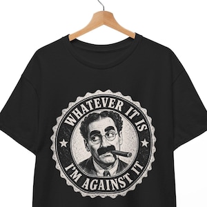 Pode incluir: Uma t-shirt preta de manga curta com uma impressão gráfica circular de um homem com bigode e um charuto. O texto "WHATEVER IT IS I'M AGAINST IT" está impresso em letras maiúsculas brancas dentro da borda circular em torno do retrato.