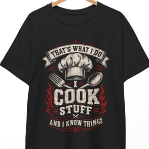 May include: A black short-sleeve t-shirt featuring a graphic print with a chef hat, a fork, and a spoon. The text reads: THAT'S WHAT I DO, I COOK STUFF, AND I KNOW THINGS. The design includes red flame accents and white lettering on a dark background.
