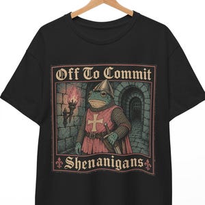 Puede incluir: Una camiseta negra con un estampado gráfico de una rana vestida de caballero medieval con cota de malla y casco, sosteniendo una espada. El texto "Off To Commit SHENANIGANS" aparece en una fuente gótica sobre e ilustracion.