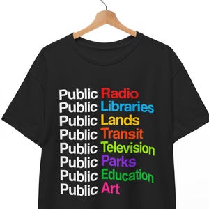 May include: A black short-sleeve t-shirt featuring the word Public in white repeated eight times, followed by the colorful words Radio in red, Libraries in blue, Lands in yellow, Transit in orange, Television in lime, Parks in purple, Education in green, and Art in pink.