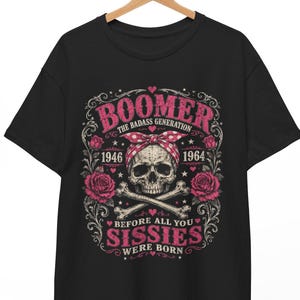 May include: A black t-shirt featuring a graphic print of a skull wearing a red and white polka dot bandana, crossed bones, and pink roses. The text reads BOOMER THE BADASS GENERATION 1946 1964 BEFORE ALL YOU SISSIES WERE BORN in pink and white.