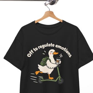 Puede incluir: Una camiseta negra con el gráfico de un pato blanco con pico y patas naranjas, que lleva una mochila verde, monta un patinete verde y sostiene una taza de café. El texto sobre el pato dice, Off to regulate emotions en color blanco.