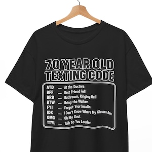 Puede incluir: Una camiseta negra de manga corta con texto blanco que dice 70 YEAR OLD TEXTING CODE seguido de una lista de abreviaturas y sus significados, incluyendo ATD At the Doctors, BFF Best Friend Fell, BRB Bathroom, Ringing Bell, entre otros.