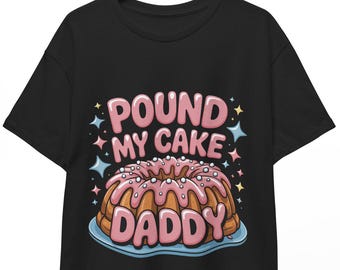 May include: Black t-shirt featuring a cartoon image of a bundt cake with pink frosting and the text "POUND MY CAKE DADDY" in pink and white. The design includes star accents.