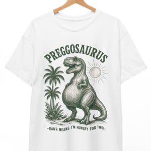 Op de afbeelding: Een wit t-shirt met korte mouwen en een donkergroene afbeelding van een dinosaurus met een dikke buik. De tekst PREGGOSAURUS staat in een boog boven de dinosaurus en de tekst -RAWR MEANS I'M HUNGRY FOR TWO!- staat onder de illustratie.
