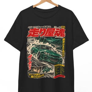 May include: A black short-sleeved t-shirt featuring a graphic print of a green sports car drifting on a mountain road. The design includes Japanese text, a portrait of a driver, and various automotive-themed illustrations in red, white, and green.
