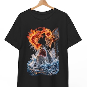 Puede incluir: Una camiseta negra de manga corta con un estampado gráfico de un tiburón emergiendo de las olas del océano hacia una canasta de baloncesto. Un camarón en llamas encesta un balón en la canasta sobre el tiburón, con llamas naranjas y rojas.