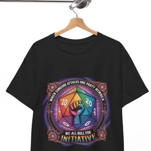 Könnte beinhalten: Schwarzes T-Shirt mit einem farbenfrohen, geometrischen Würfeldesign und dem Text "WHEN SOMEONE ATTACKS ONE PARTY MEMBER WE ALL ROLL FOR INITIATIVE." Das Design beinhaltet eine Faust und die Zahlen 20, 6 und 8. Das Shirt hängt an einem Holzbügel.