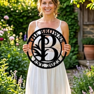 Peut inclure: Panneau de nom de famille en métal noir avec l'inscription "THE BROWNS" et "EST 2023" encerclant un grand "B" avec un motif de branche décoratif. Le panneau rond est tenu devant une personne dans un jardin.