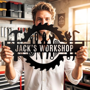 Puede incluir: Un letrero de metal negro con la silueta de una hoja de sierra circular y formas de herramientas integradas como un martillo, alicates, llave inglesa y destornillador, con el texto JACK'S WORKSHOP en letras blancas en el centro.