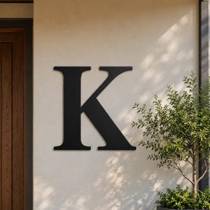 Puede incluir: Una letra K grande de metal negro mate montada en una pared exterior de color claro. La letra presenta una tipografía clásica con remates y bordes limpios y definidos, situada a la derecha de una puerta de madera oscura.